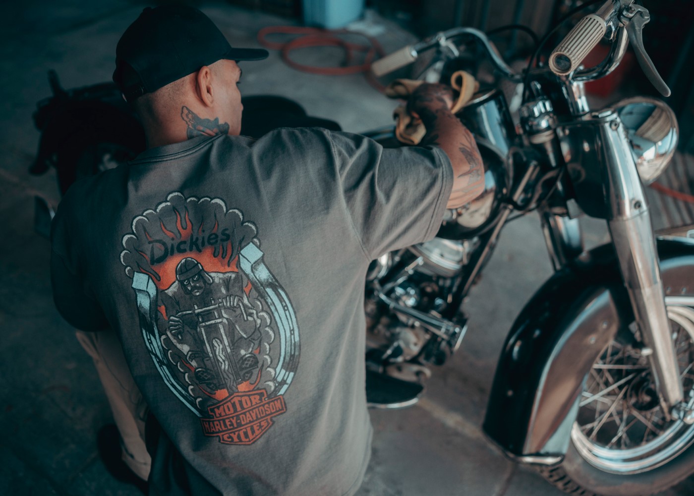 A man wearing a graphic t-shirt with Dickies and Harley-Davidson logos on backside crouched next to motorcycle