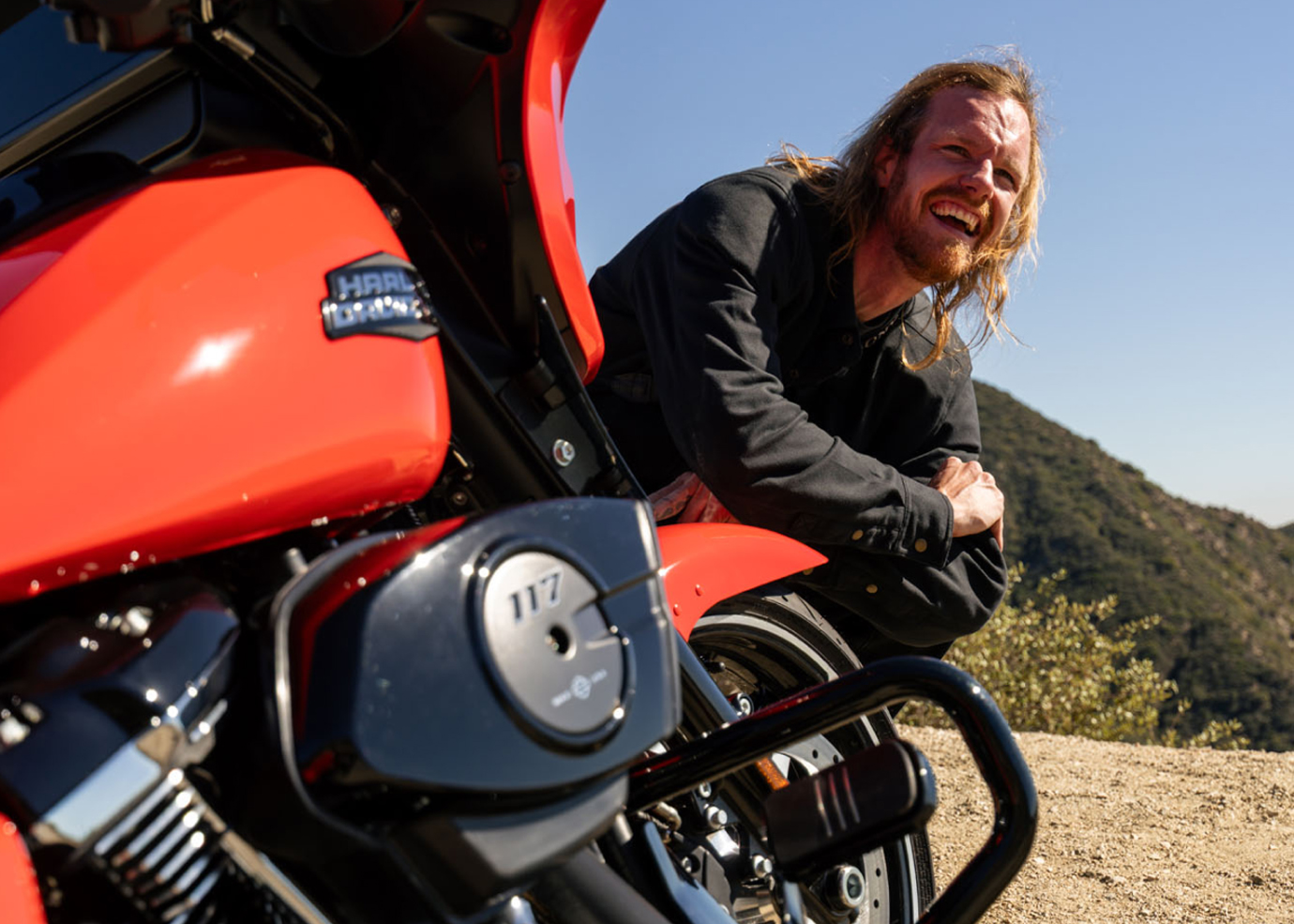 A person leaning against a red HD motorcycle at a dirt overlook